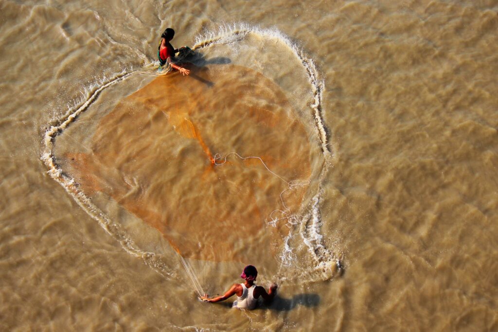 Drone view of anonymous ethnic female catching fish with net in brown lake with rippled water in daylight